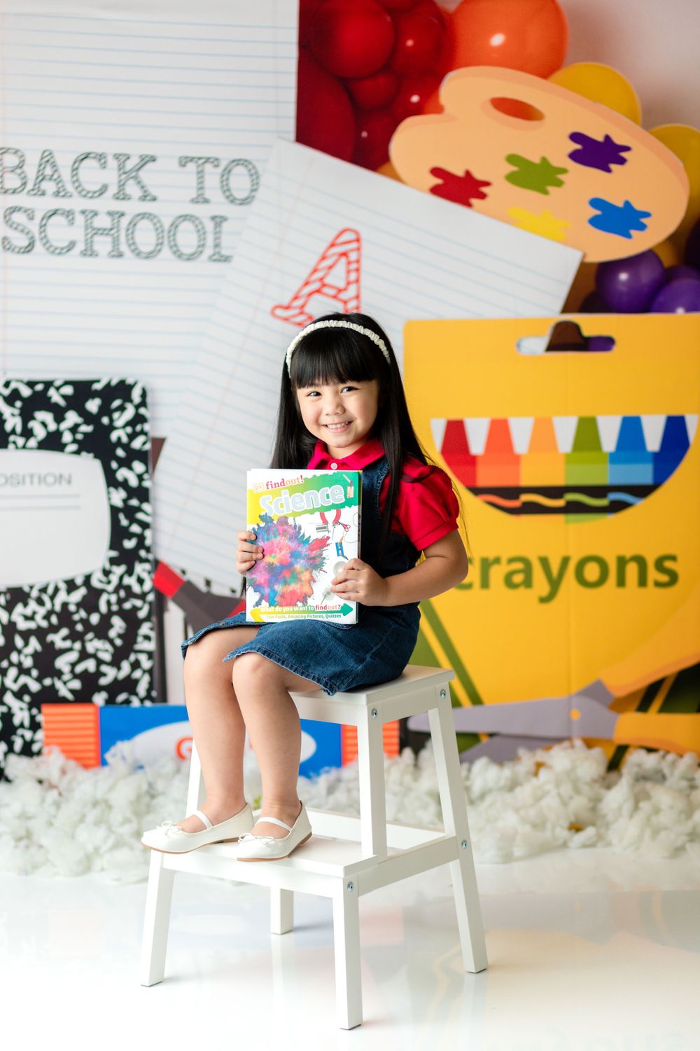 A student in denim overalls sits on a white stool holding a colorful book against school-themed decorations.