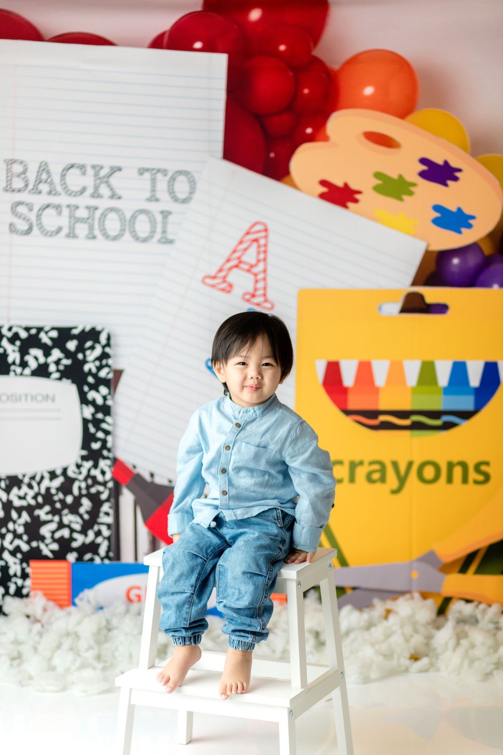 A student in light blue attire sits on a white stool against school-themed decorations and red balloons.