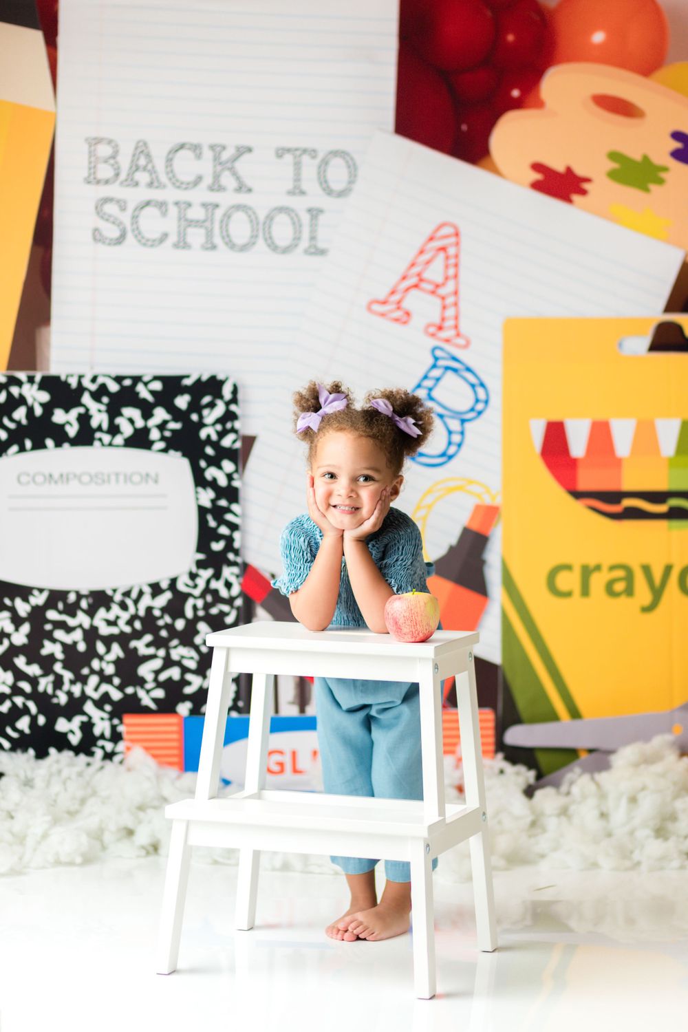 A student poses cheerfully at a white desk with back to school decorations and a giant crayon box backdrop.
