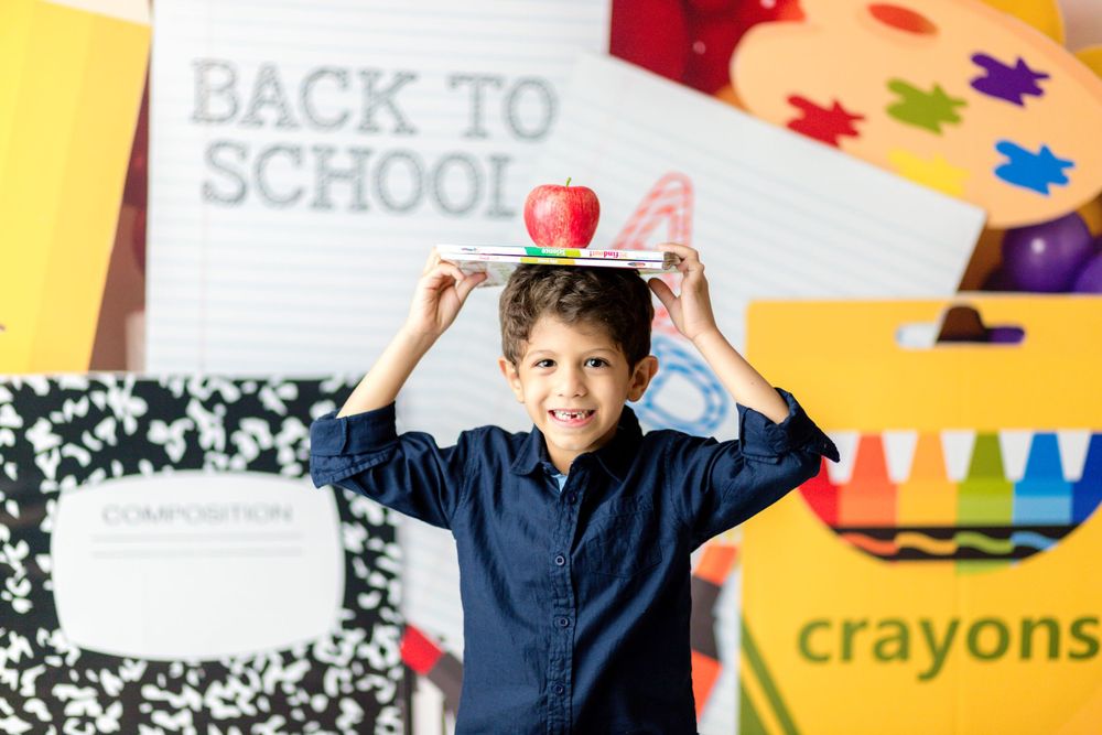 A student balances an apple on their head against a backdrop with composition notebooks and art supplies.