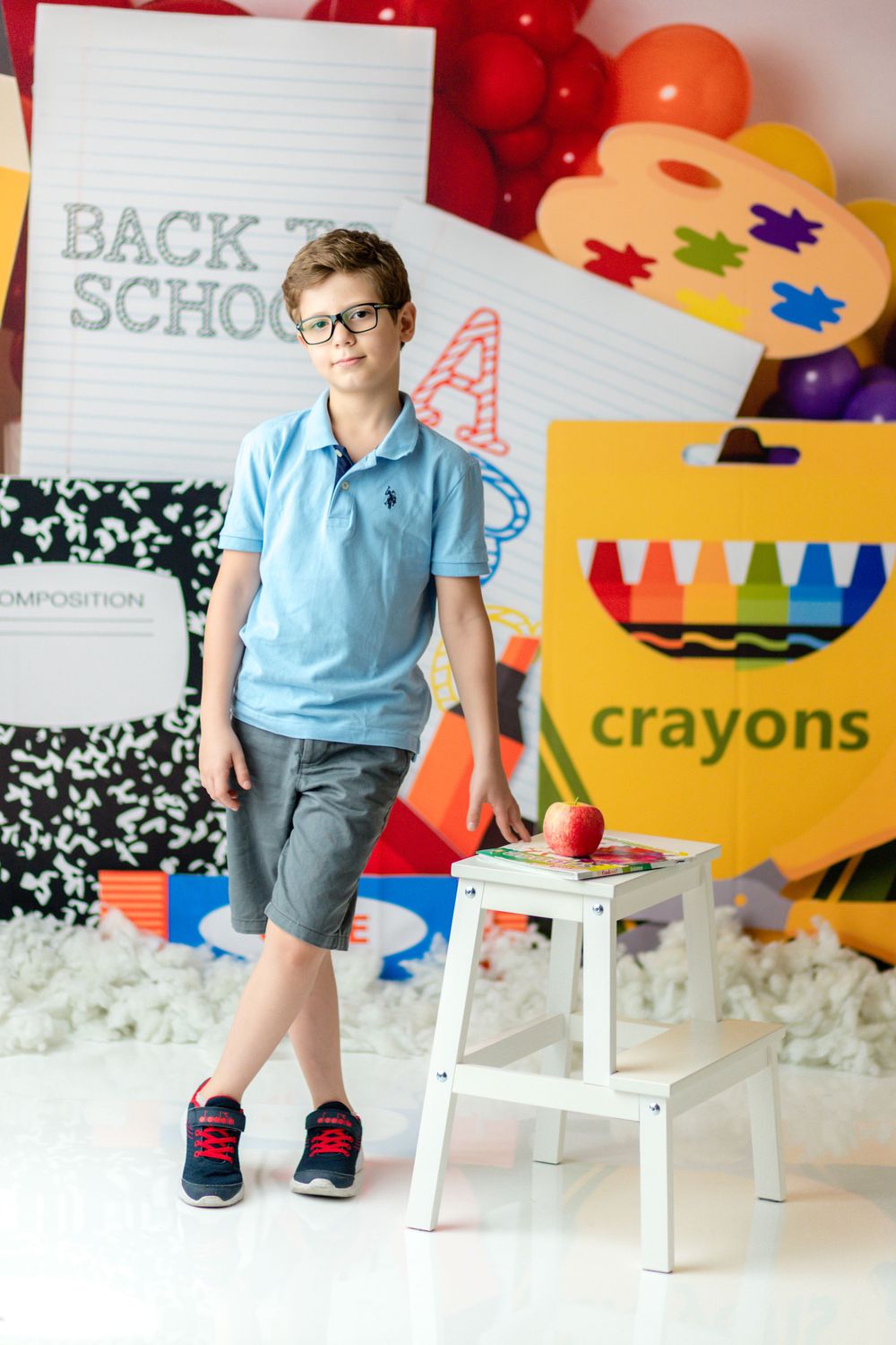 A student in a blue polo shirt stands next to a white stool against a colorful back-to-school backdrop.