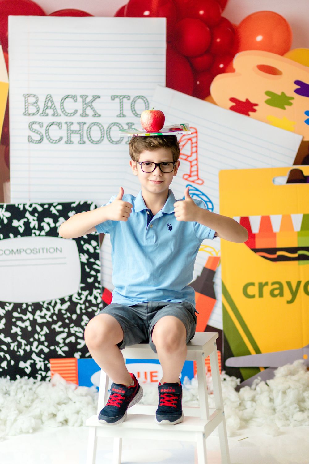 A student gives thumbs up while sitting on a white stool with apples and school-themed decorations.