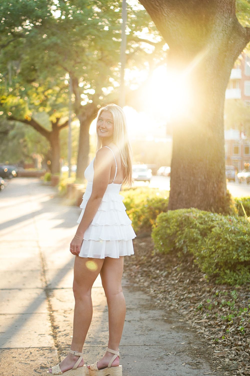Sunlit portrait of someone in a white dress standing on a tree-lined path at golden hour.