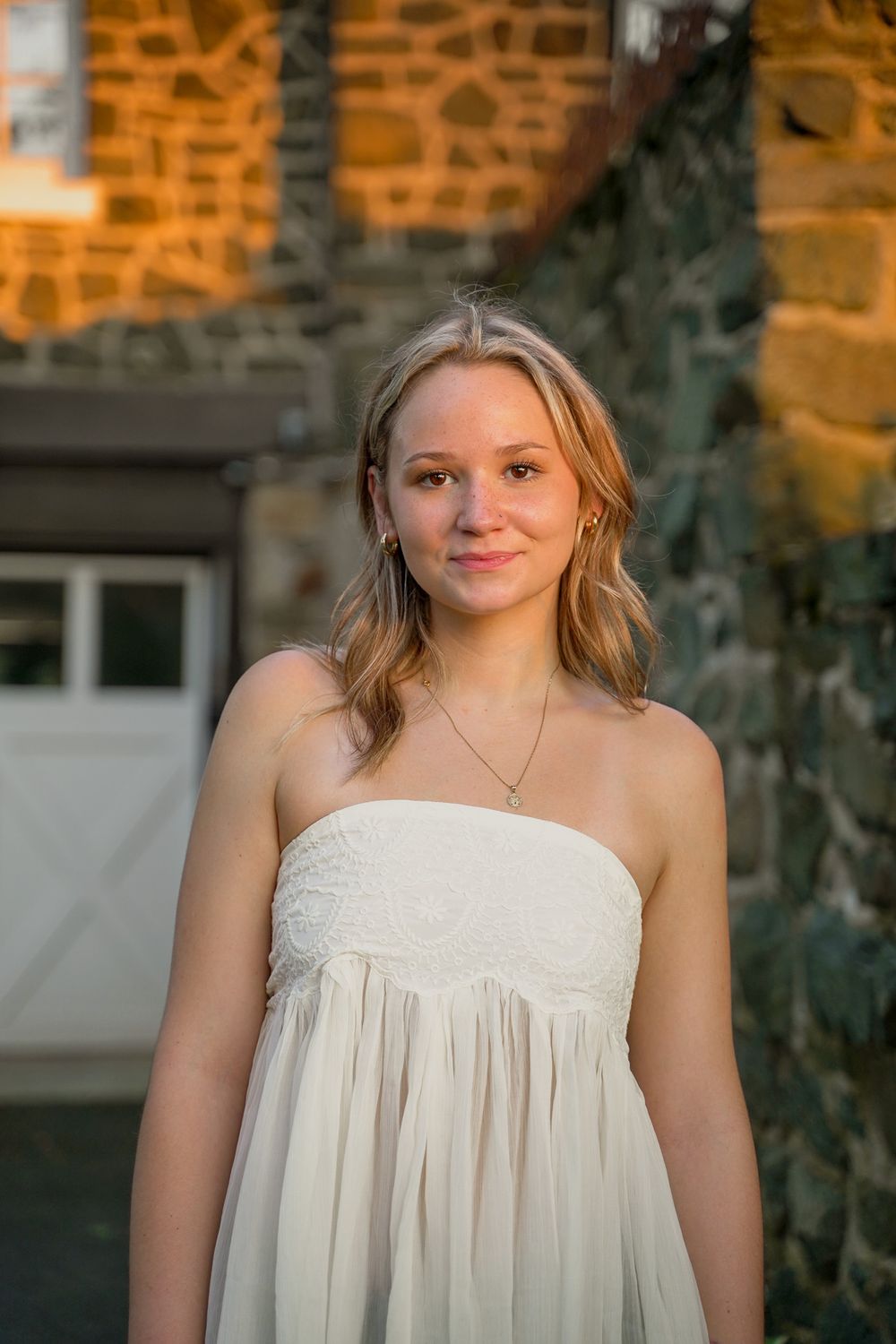 Portrait of young woman in white strapless dress against stone wall at sunset.