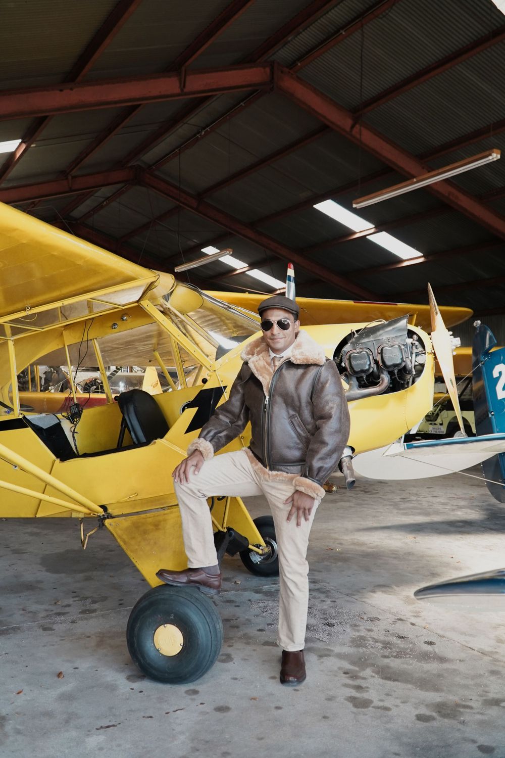 Vintage yellow aircraft in hangar with person posing beside it in aviation attire.