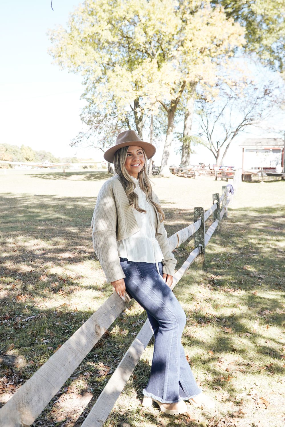 Casual western-inspired outfit featuring denim and hat while leaning on rustic wooden fence.