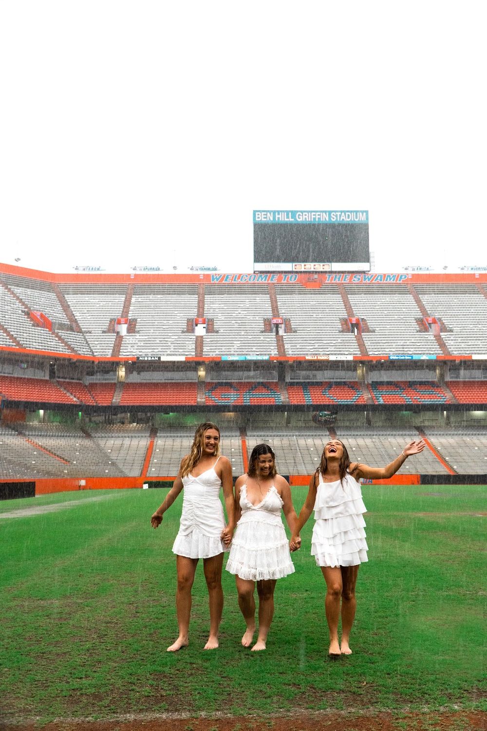 Three friends in white dresses celebrate on football field in front of stadium seating. Graduation