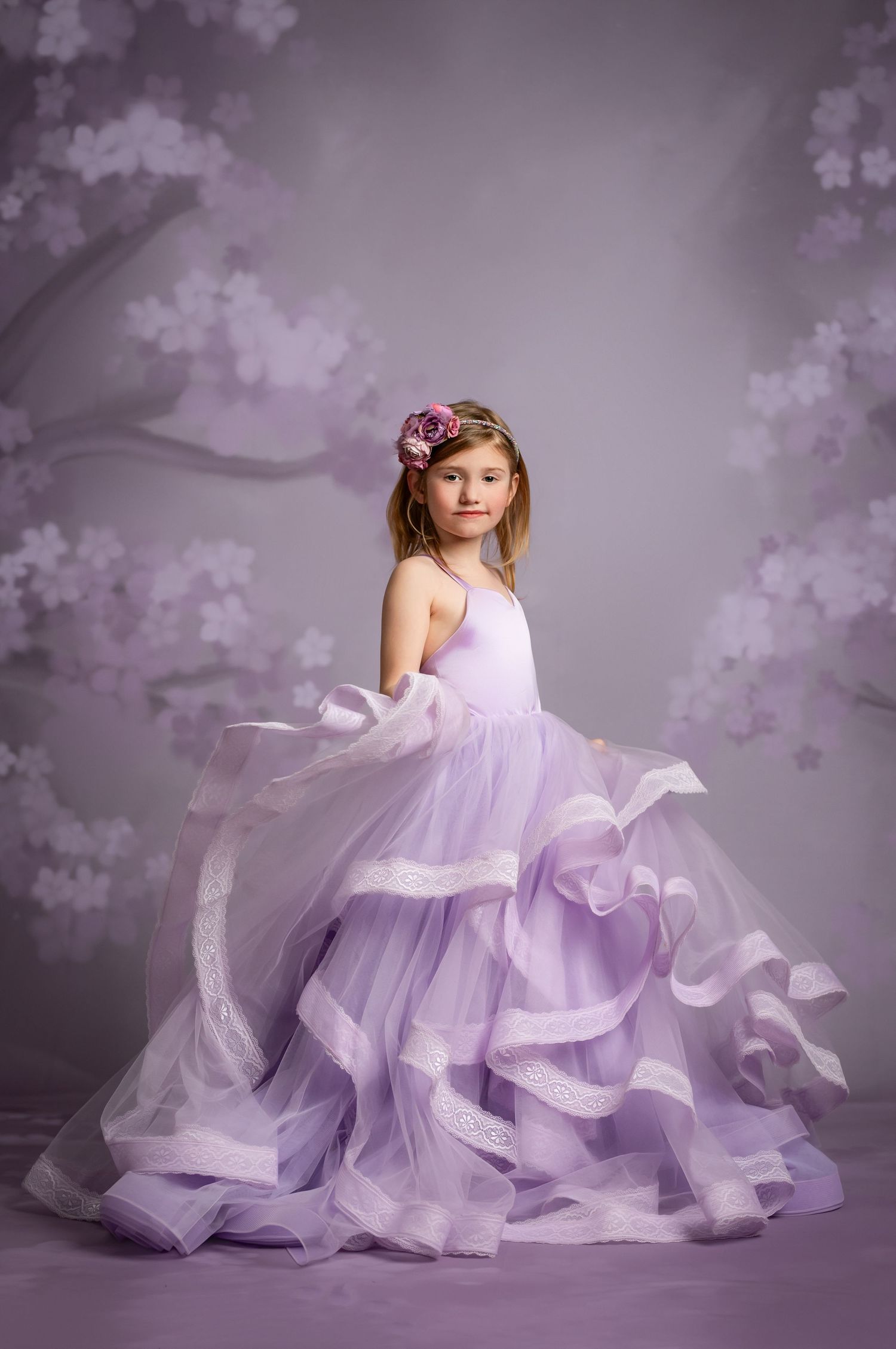 A flowing lavender ballgown with ruffled layers twirls gracefully in a dreamy purple-tinted studio setting with floral backdrop.