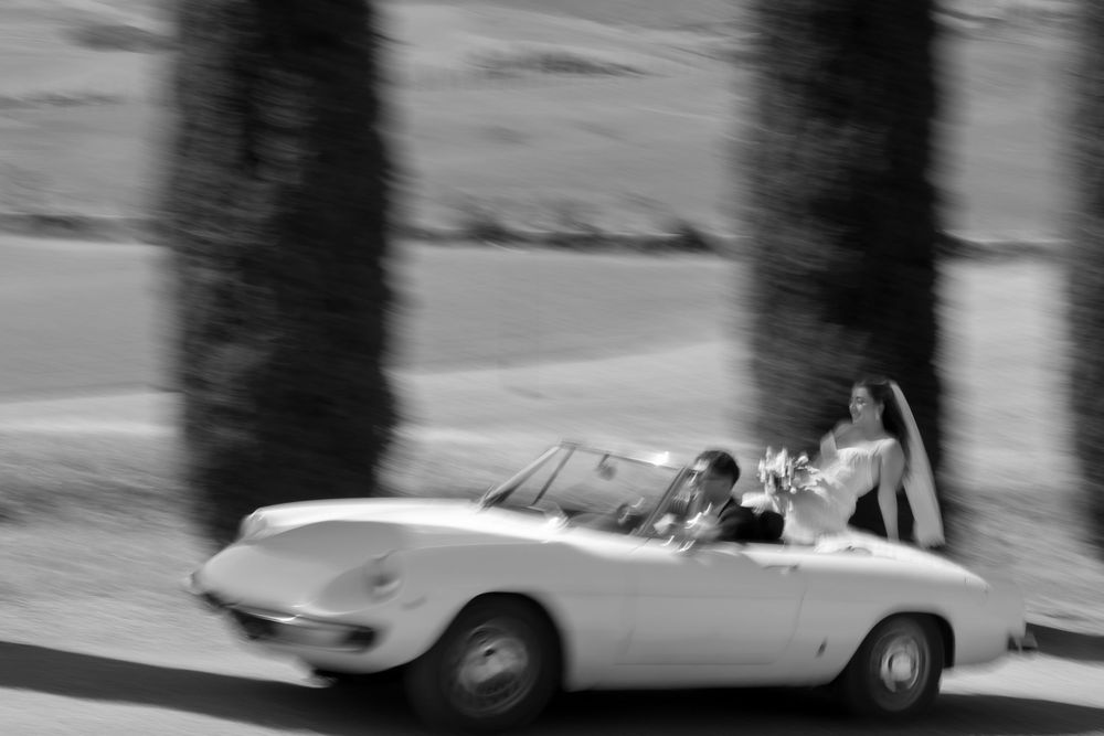 Black and white photo of a bride and groom riding in a classic white convertible sports car on their wedding day.