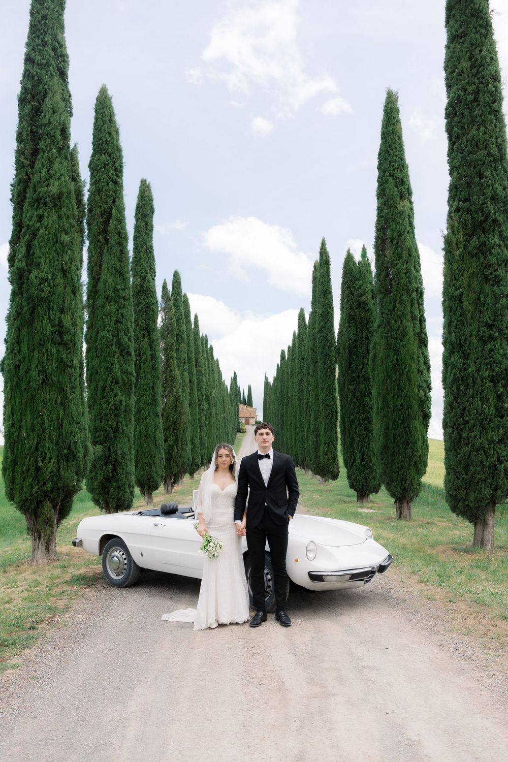 Bride and groom posing by a white vintage convertible on a cypress tree-lined gravel path in Tuscany.