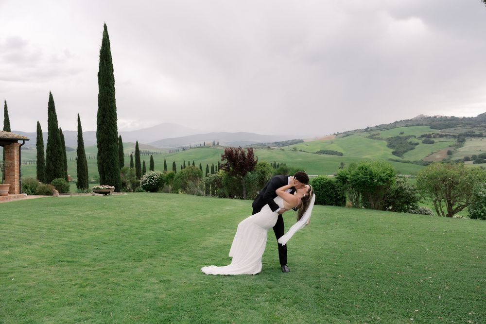 Bride and groom share a romantic dip kiss on lush green lawn with Tuscan cypress trees and rolling hills backdrop.