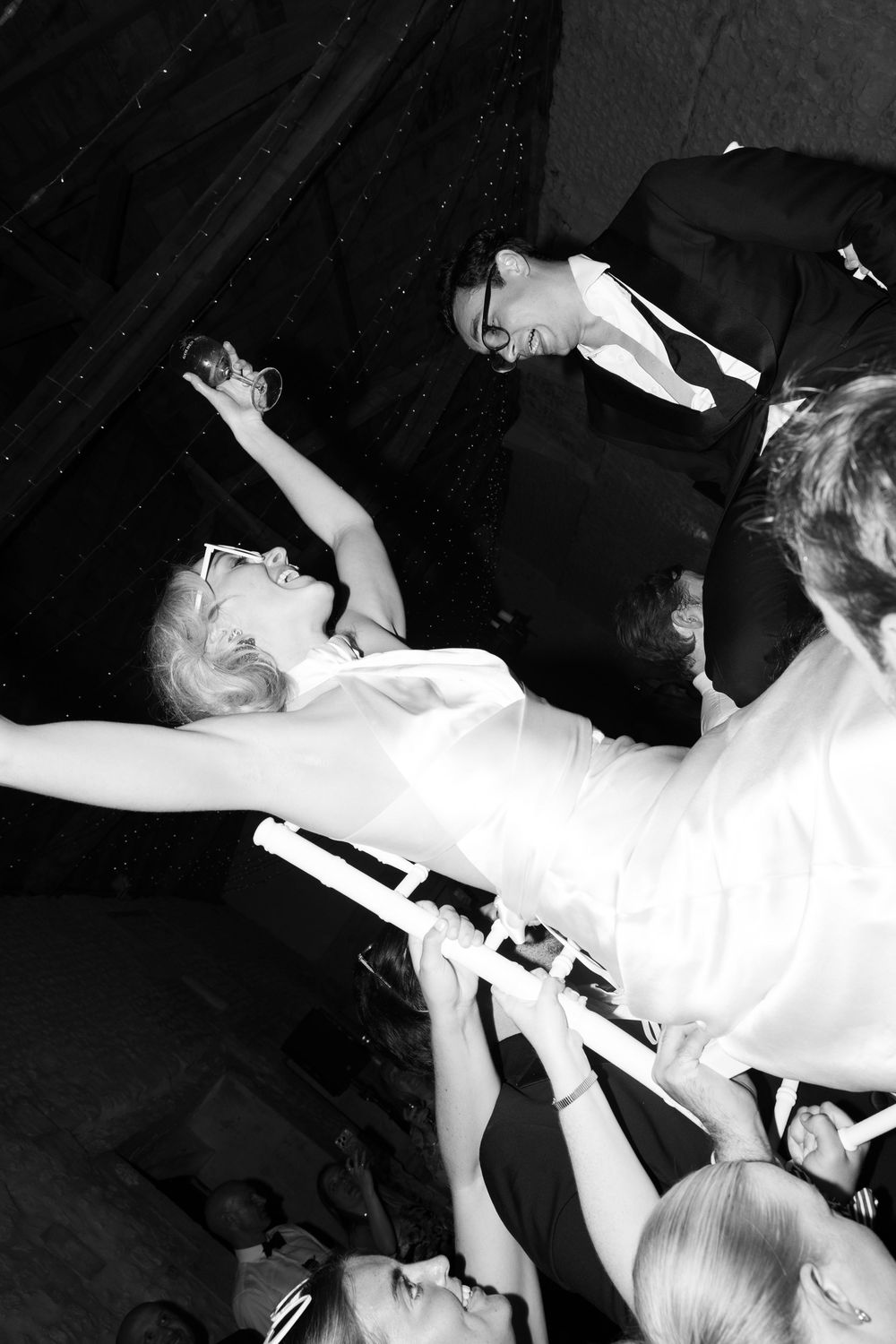Black and white photo of a bride being lifted by guests at a lively wedding reception celebration.