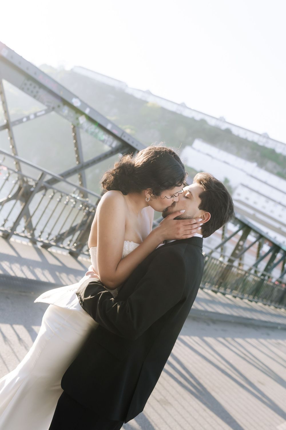 Bride and groom share a romantic kiss on a bridge, with the groom lifting his bride in a white strapless gown.