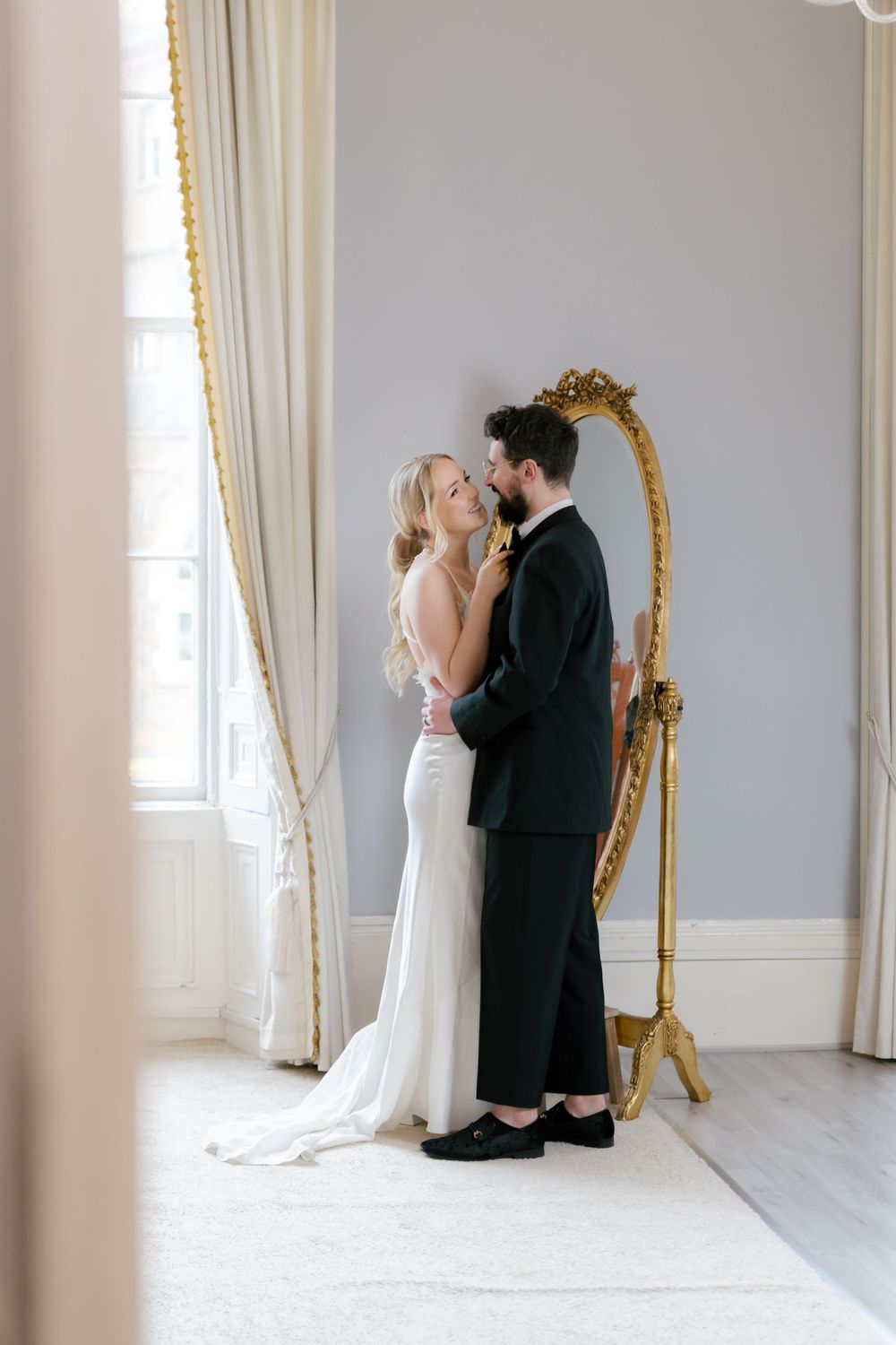 A romantic wedding moment captured in an elegant white room with a gilded mirror and soft natural light.