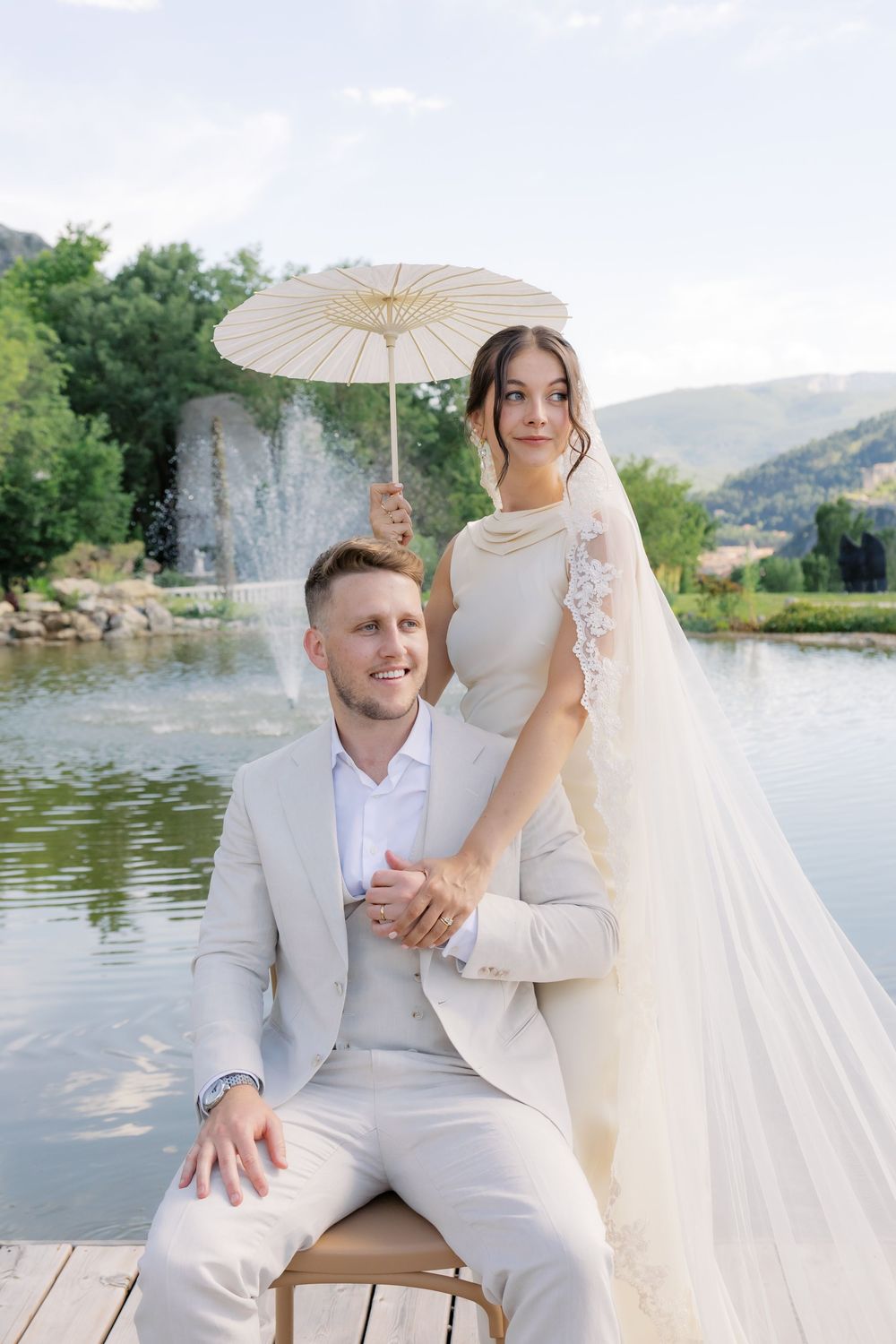 Elegant bride holding white parasol stands behind smiling groom in beige suit on dock with fountain and mountains backdrop.