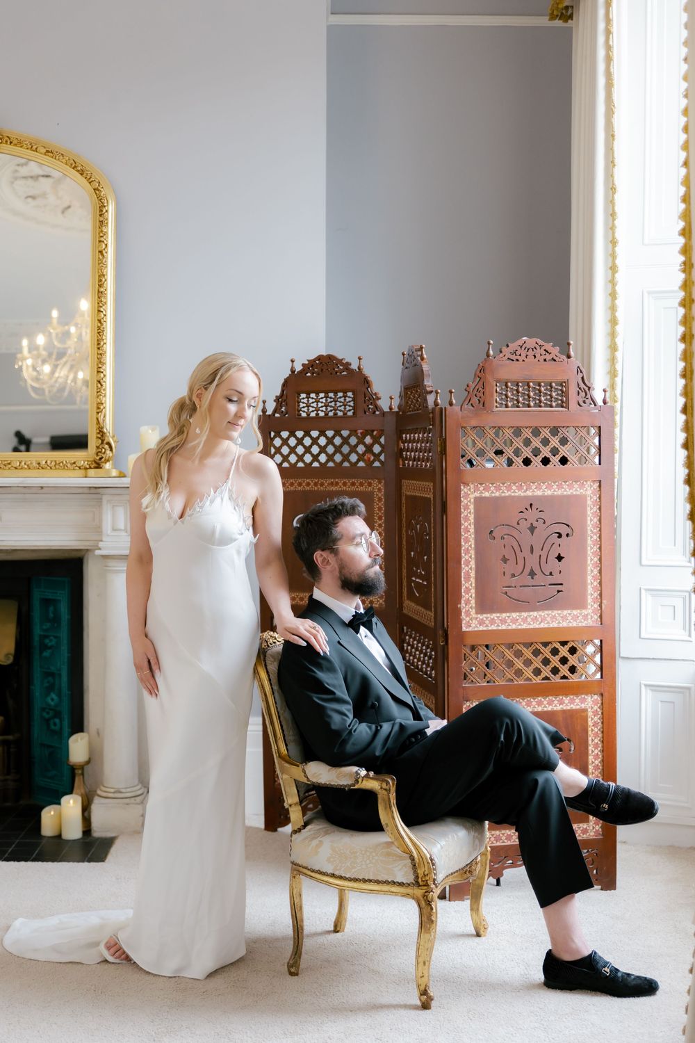 Wedding couple poses casually against vintage wooden screens and gold-framed mirrors in a bright room.