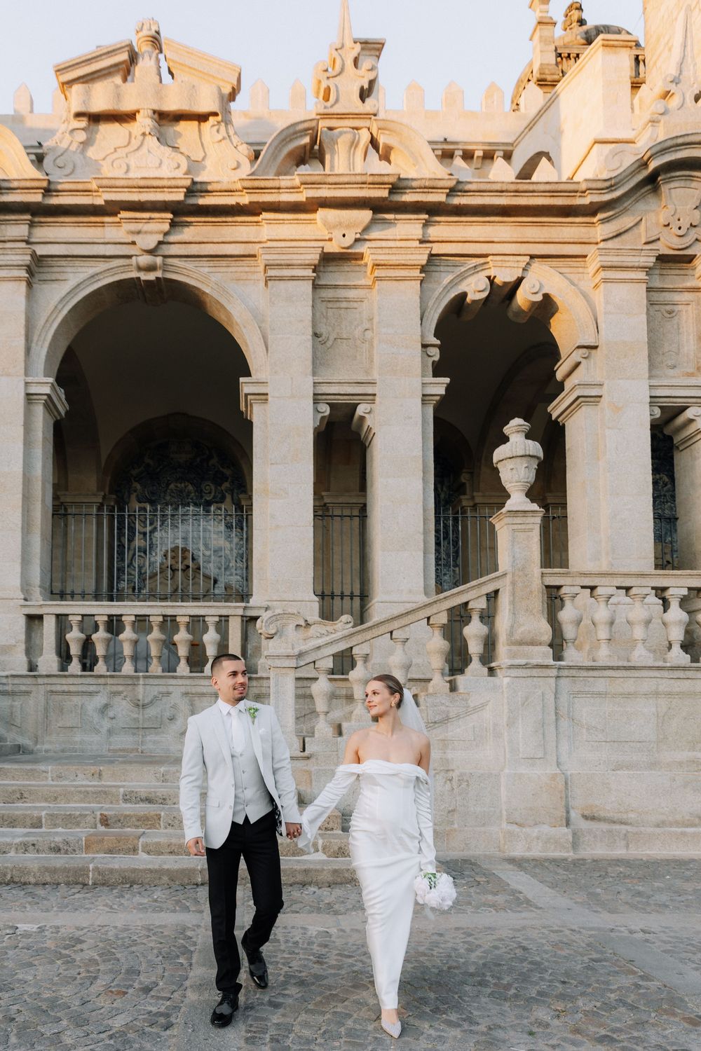 Couple in formal white attire runs playfully through ornate stone courtyard with classical architecture.