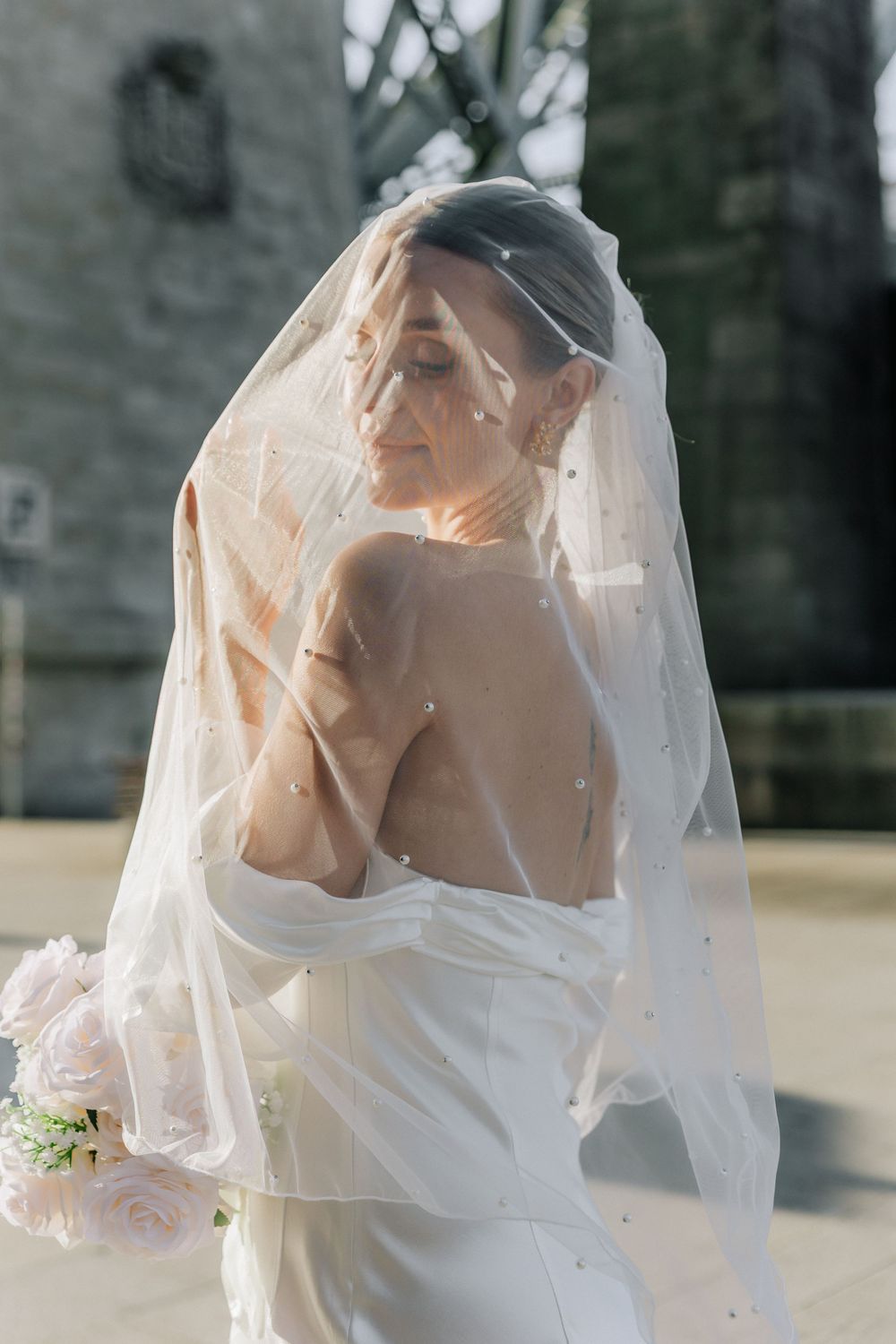 Elegant bride in white off-shoulder gown and delicate veil poses with soft sunlight creating a romantic bridal portrait.