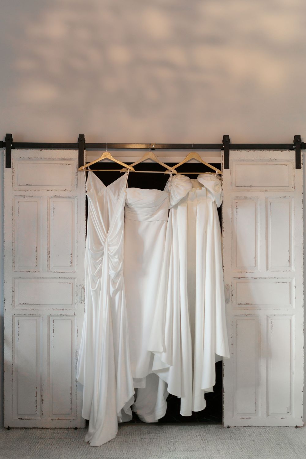 Three elegant white wedding dresses hanging on black hangers against rustic white wooden doors.