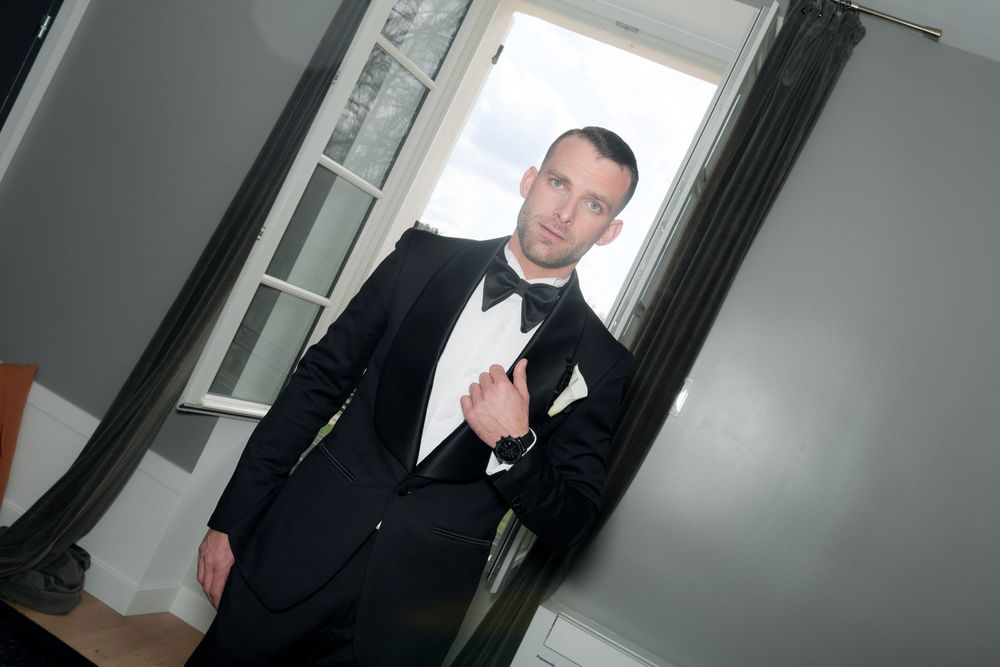 A person in a black tuxedo with bowtie stands in a doorway, adjusting their jacket.