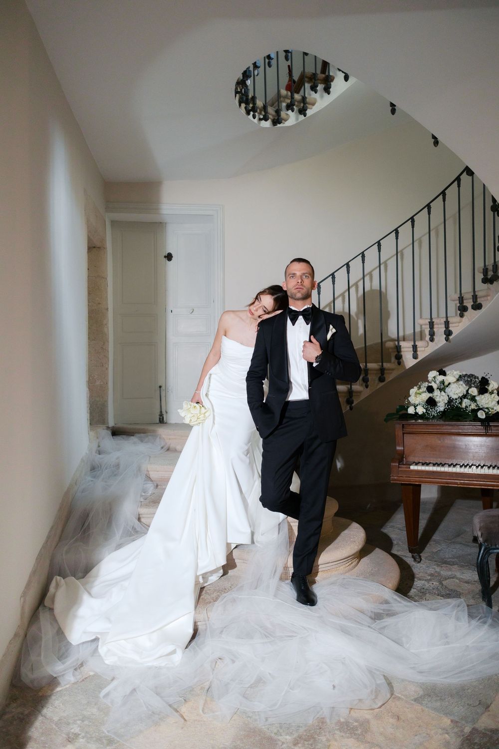A couple in formal wedding attire pose on elegant marble stairs near a piano in an indoor setting.