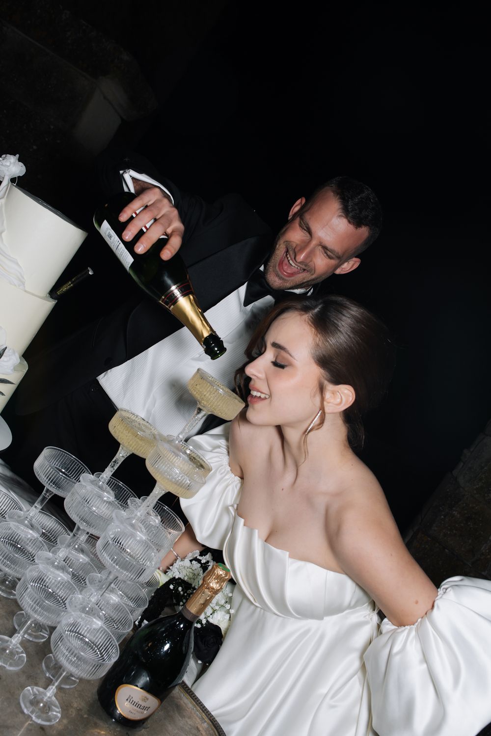 A couple pours champagne into a tower of crystal glasses at an elegant wedding celebration.