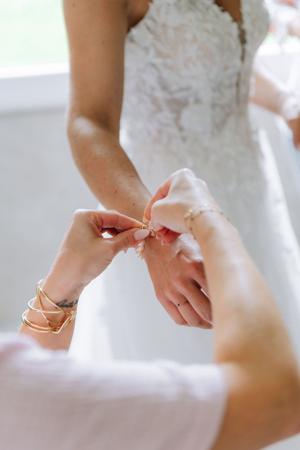 A close-up of hands fastening a delicate pearl bracelet against the backdrop of a white lace wedding dress.