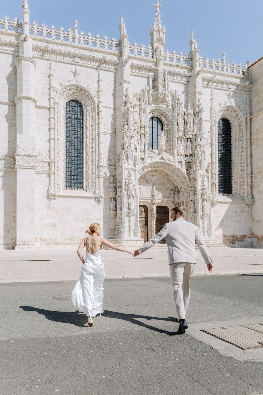 A couple holding hands runs towards an ornate white Gothic church with tall arched windows and decorative spires.