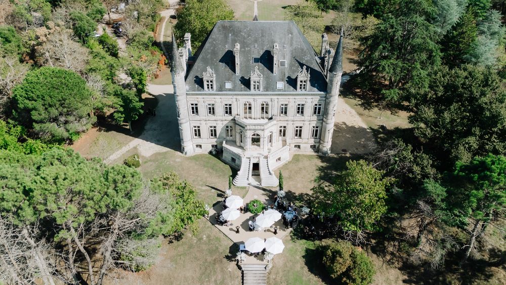 Aerial view of a grand white mansion with multiple chimneys surrounded by lush green trees and landscaping.