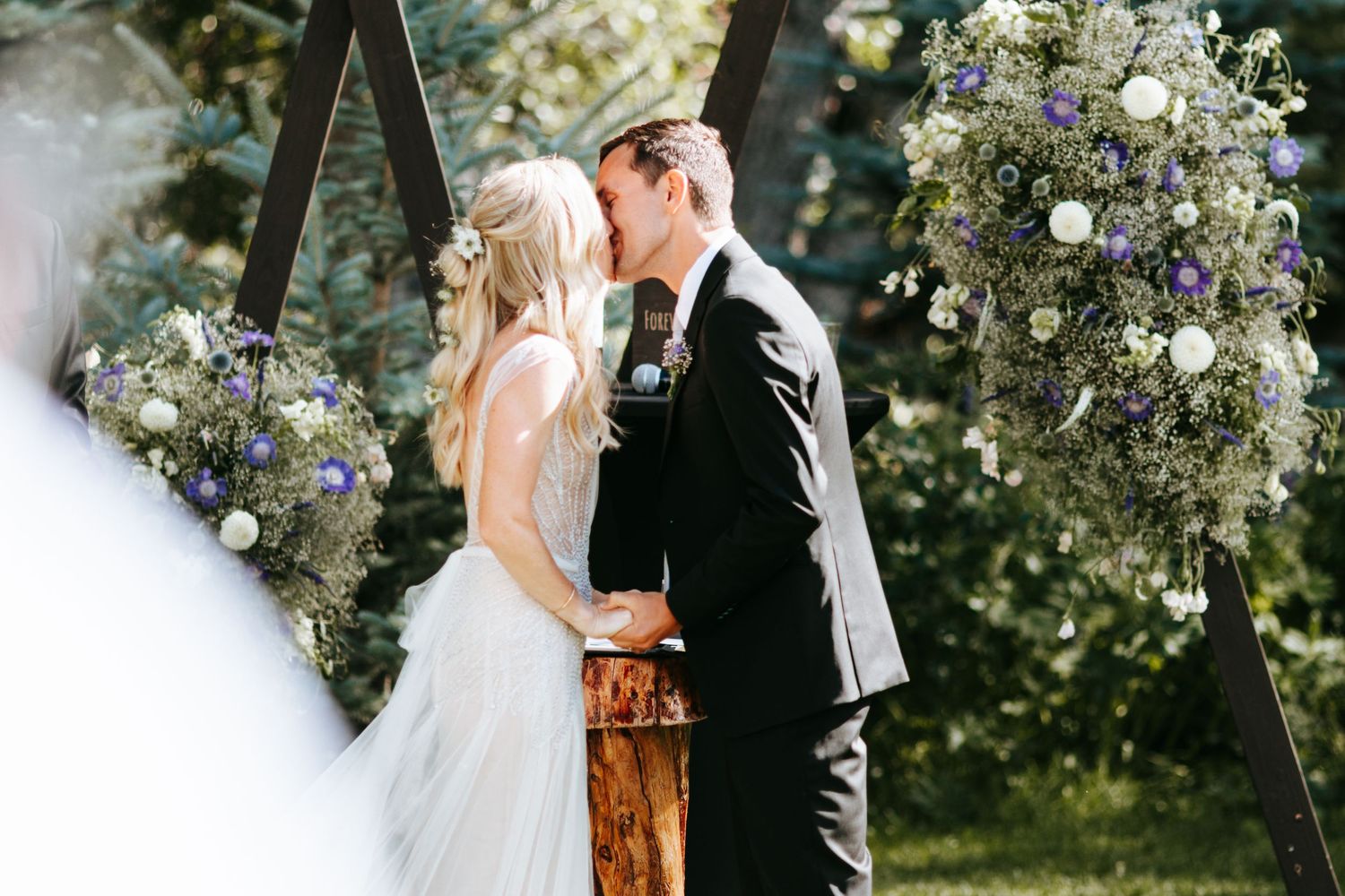 A romantic outdoor wedding ceremony under an arch decorated with white and lavender flowers in a garden setting.