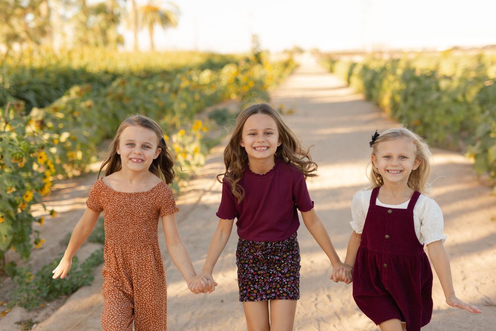 Three children holding hands while walking down a path surrounded by sunflowers.