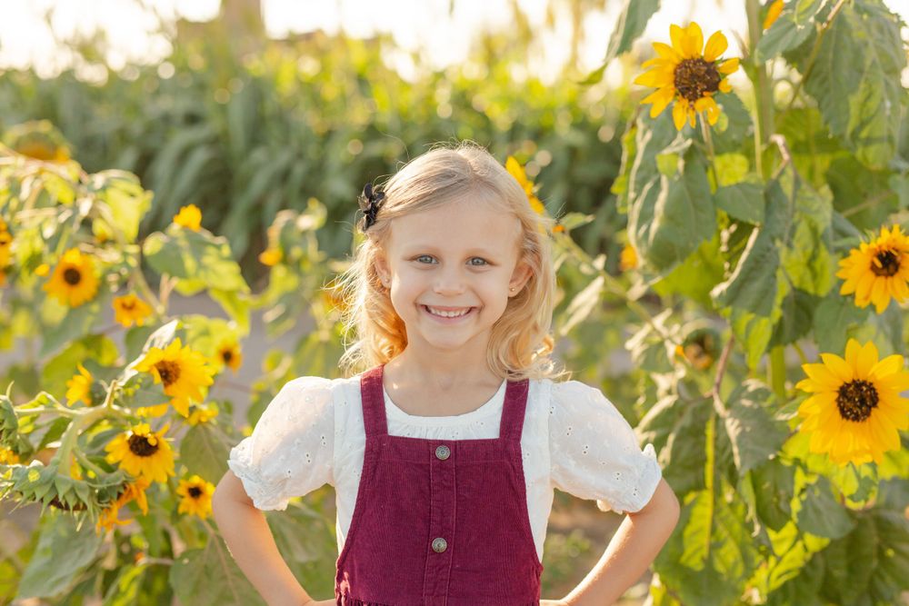 Young child wearing burgundy overalls and white top poses cheerfully in a blooming sunflower field at golden hour.