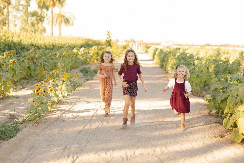 Three children run joyfully down a path lined with bright yellow sunflowers.