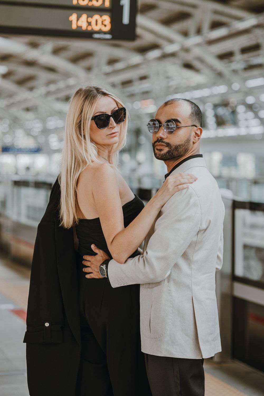Stylish couple in sunglasses posing at a train station, woman in black dress and man in white blazer embracing.