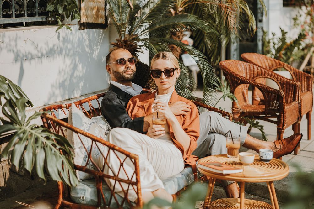 Stylish couple in sunglasses relaxing on rattan furniture surrounded by tropical plants on a sunny patio.