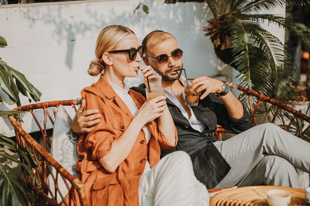 Stylish couple sitting in rattan chairs sipping drinks outdoors surrounded by tropical plants.