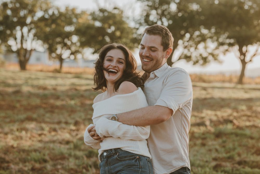 Happy couple embracing and laughing together in a golden sunlit field with trees in the background.