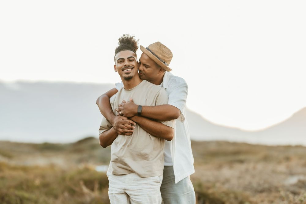 Two young Black men share a tender embrace outdoors, one kissing the other on the cheek while smiling.