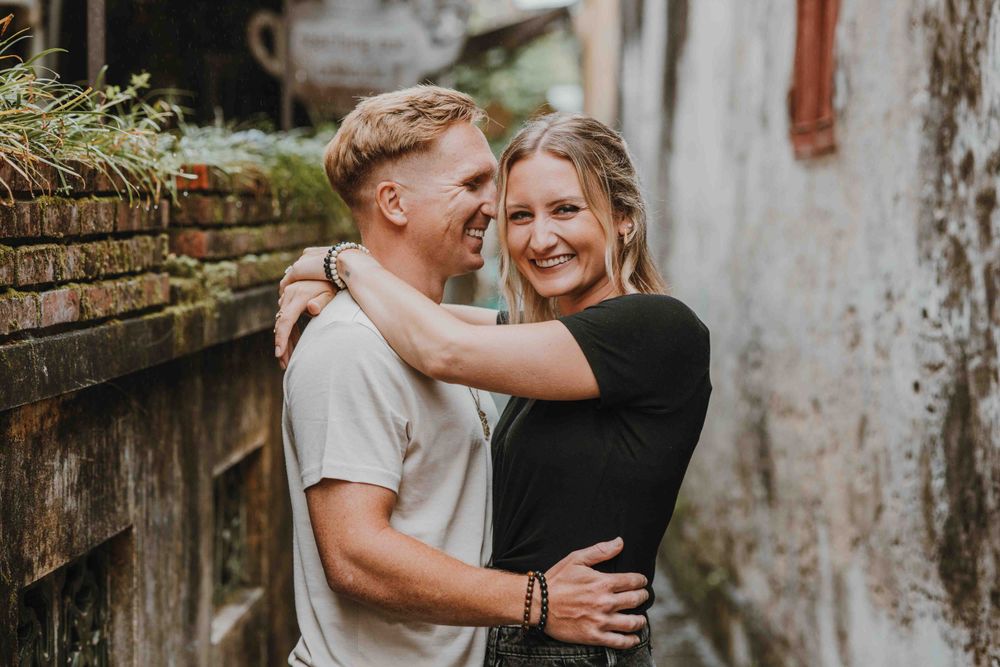 A happy couple embraces and laughs together against a weathered wall with rustic brick details.