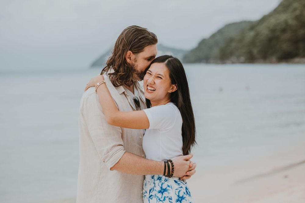 Couple embracing and laughing on a tropical beach with lush green hills and calm ocean in the background.