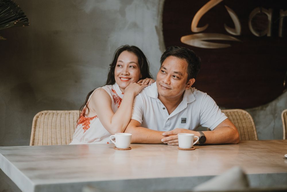 A smiling couple sits together at a cafe table with white coffee cups, posing for a romantic portrait photo.