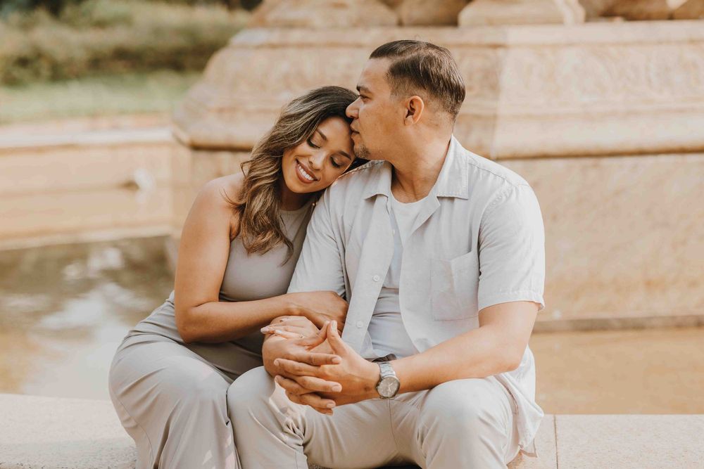 A couple sits together outdoors, sharing a tender kiss near a fountain, dressed in matching neutral tones.