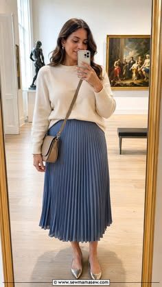 Woman in cream sweater and blue pleated midi skirt takes mirror selfie, styled with silver flats and tan crossbody bag.