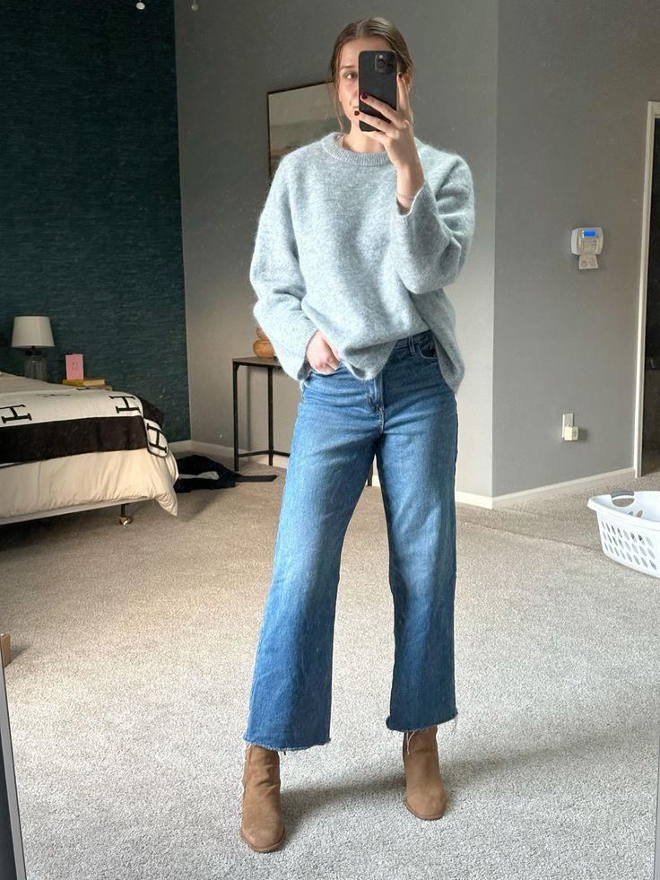 Woman in light gray sweater, wide-leg cropped jeans, and tan ankle boots taking a mirror selfie in a bedroom.