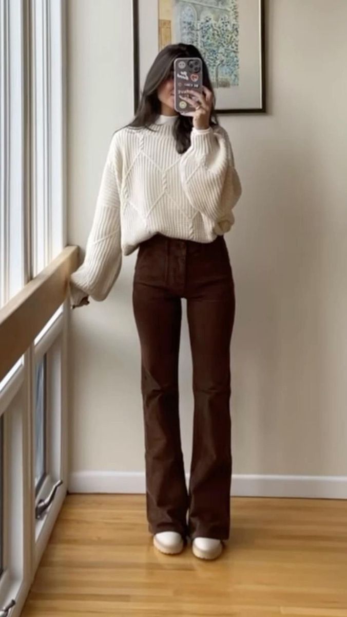 Woman wearing a cream sweater and brown wide-leg pants poses by a window in a neutral toned room with hardwood floors.
