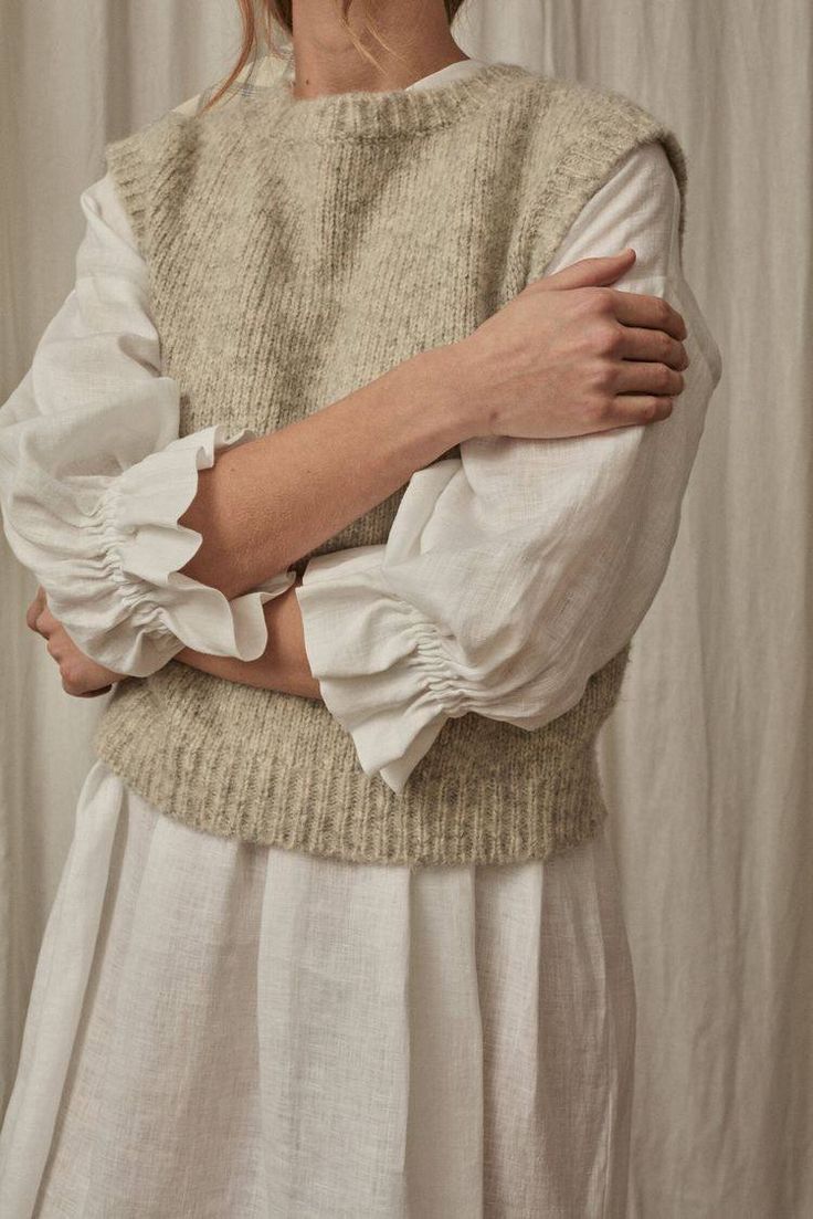 Beige knit vest styled over white puff sleeve dress against neutral background.