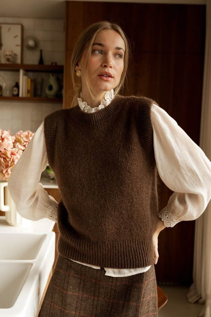 Woman wearing a brown knit vest over a white lace-collar blouse, paired with a plaid skirt, standing in a cozy kitchen.