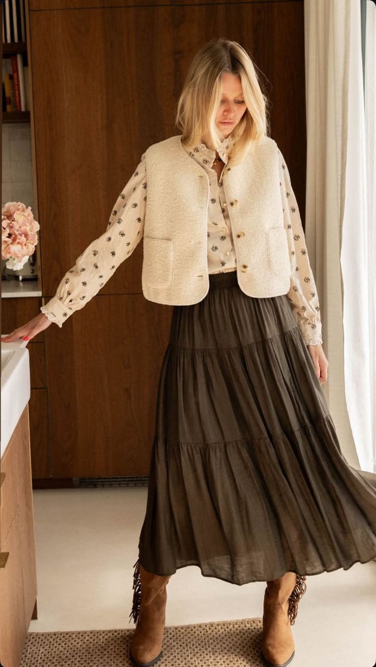 Woman wearing cream tweed vest over floral blouse with dark olive maxi skirt and fringe ankle boots.