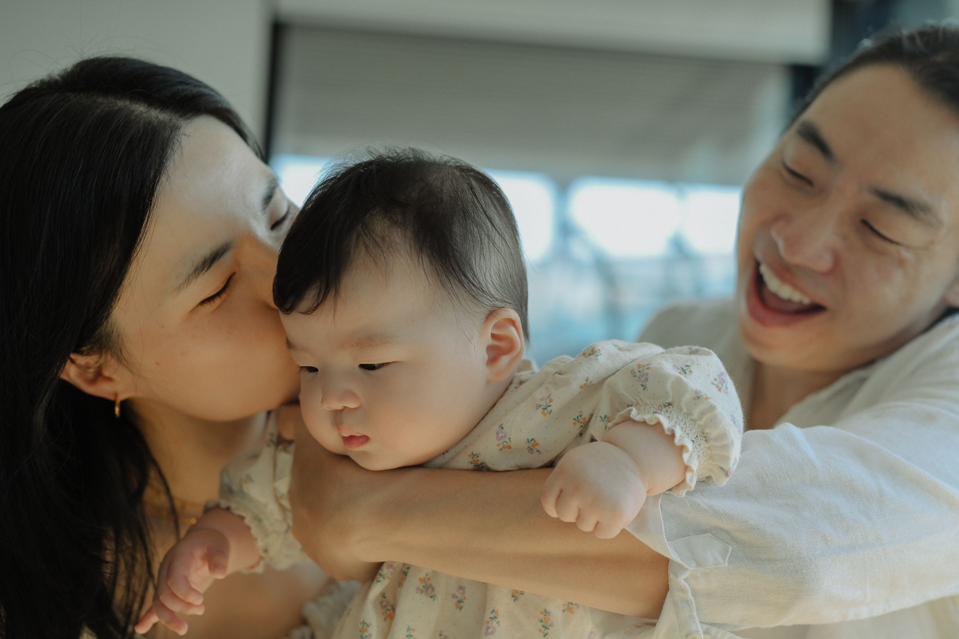 A tender family moment as parents share loving affection with their baby in soft natural lighting.