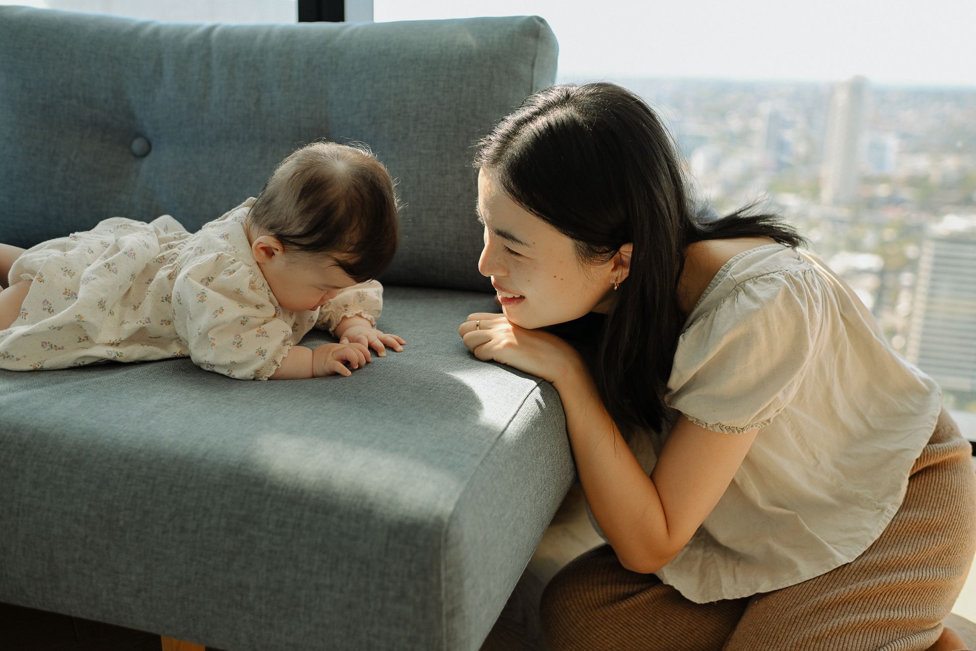 A person in a white top leans on a gray couch while playing with a small dog in a bright room with city views.